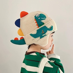 May include: A cream-colored knit beanie featuring a green dinosaur graphic on the side. The hat includes a ribbed texture and colorful, protruding fabric spikes in orange, blue, and yellow along the back, extending into a tail-like shape.