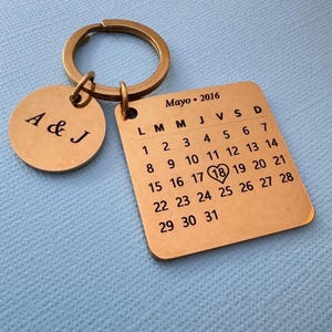 Può includere: Un portachiavi dorato con un piccolo tag rotondo inciso con "A & J" e un tag rettangolare più grande con un calendario per maggio 2016. Il numero 18 è evidenziato con un cuore.