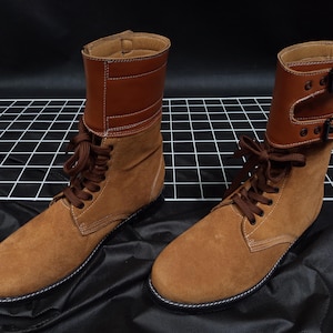 Könnte beinhalten: Ein Paar braune Wildlederstiefel mit Lederakzenten. Die Stiefel haben ein Schnürdesign und eine hohe Silhouette. Der obere Teil der Stiefel weist dunkleres braunes Leder mit Schnallendetails auf. Die Sohlen sind hellbraun mit weißen Nähten.
