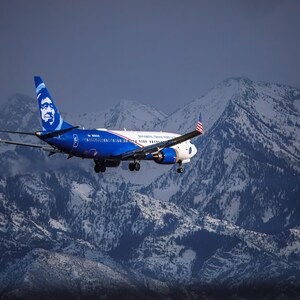 May include: An Alaska Airlines jet, predominantly blue and white, flying over snow-capped mountains. The aircraft features the airline's logo on the tail and wings, with the American flag visible on the wingtips.