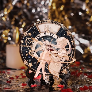 May include: A decorative clock with a celestial design and a cartoon character. The clock face is dark blue with gold accents, stars, and Roman numerals. The character is in a white dress. The clock is displayed on a black stand.