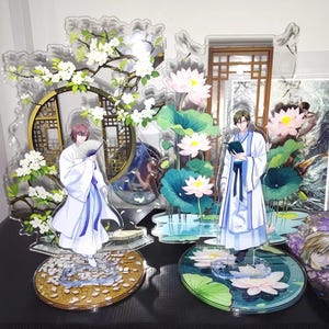 以下が含まれることがあります： 白いローブと青いアクセントを身に着けた2人のキャラクターが特徴のアクリルアニメフィギュア。一人は扇子を、もう一人は本を持っており、桜、蓮の花、伝統的なアジアの建築要素を背景にしています。