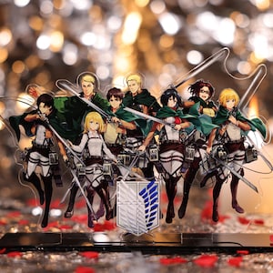 Op de afbeelding: Een heldere acrylstandaard met een groep anime-personages in actieposes, elk met een zwaard. De personages dragen witte, bruine en groene uniformen. Een wapenschild met blauwe vleugels staat aan de basis.