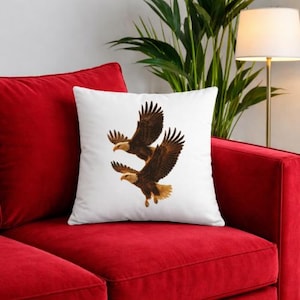 May include: White square cushion featuring a detailed illustration of two bald eagles in flight. The eagles have brown and white feathers, with outstretched wings. The cushion is placed on a red sofa.