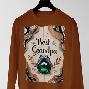 May include: A brown long-sleeved top with the text "Best Grandpa" in a decorative graphic. The graphic includes a sea lion, a large green gem, and decorative elements. The top is displayed on a black hanger.