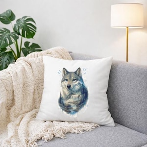 Könnte beinhalten: Quadratisches weißes Kissen mit einer Aquarellillustration eines Wolfes mit blauen Augen. Das Kunstwerk zeigt eine Berglandschaft mit Wasserfall und Bäumen. Das Kissen liegt auf einem grauen Sofa mit einer cremefarbenen Decke.