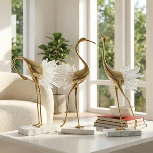 May include: Three gold-colored crane sculptures with crystal-like accents. Each crane stands on a white marble base. The sculptures are arranged on a white table, with a neutral-toned background.