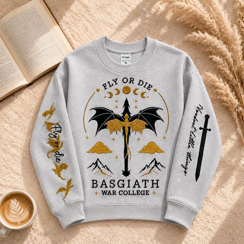Pode incluir: Um moletom cinza claro com um desenho de drag&atilde;o e o texto "FLY OR DIE" e "BASGIATH + WAR COLLEGE +". O design inclui um drag&atilde;o, luas, nuvens e montanhas em preto e dourado. A manga tem o texto "Fly or die" e "Voudost Little things".