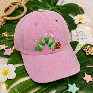 Könnte beinhalten: Rosa Baseballkappe mit den gestickten Worten "little grower" und einem bunten Raupenmuster. Die Kappe hat einen gebogenen Schirm und einen verstellbaren Riemen. Hergestellt aus einem weichen, strukturierten Stoff.