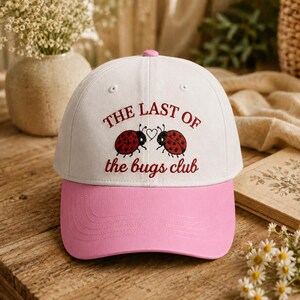 May include: White baseball cap with a pink peak. The cap features the text "THE LAST OF" above two ladybug illustrations and the text "the bugs club". The cap has a pink button on top.