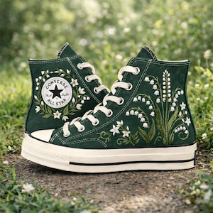 Puede incluir: Un par de zapatillas altas verde oscuro con cordones y suelas blancas. Las zapatillas presentan diseños florales bordados, incluyendo una corona alrededor del logo Converse All Star y lirios de los valles. Las zapatillas están sobre un fondo de hierba verde.