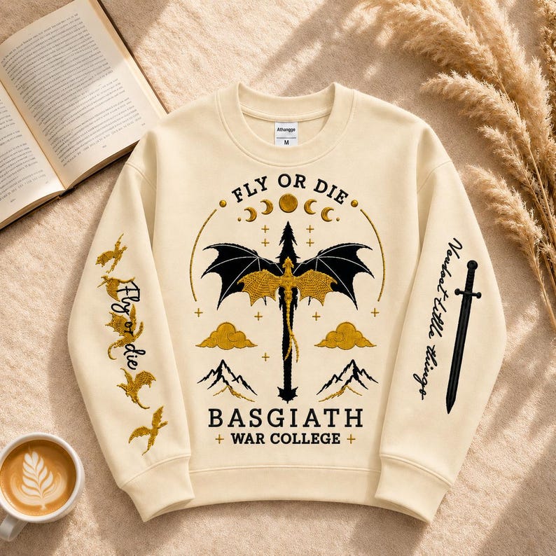 Pode incluir: Camisola creme com um desenho de drag&atilde;o e o texto "FLY OR DIE" e "BASGIATH + WAR COLLEGE +". As mangas apresentam texto adicional e ilustra&ccedil;&otilde;es de drag&otilde;es e uma espada.