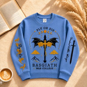Pode incluir: Um moletom azul claro com um desenho de drag&atilde;o e o texto "FLY OR DIE" e "BASGIATH + WAR COLLEGE +". As mangas t&ecirc;m texto adicional e gr&aacute;ficos de p&aacute;ssaros. Uma x&iacute;cara de caf&eacute; e um livro aberto est&atilde;o no fundo.