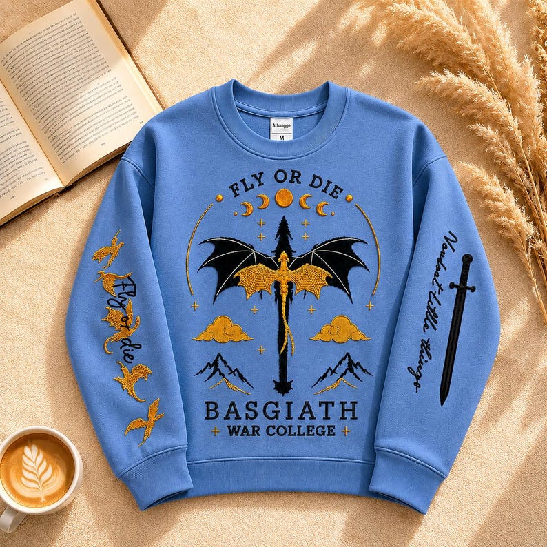 Pode incluir: Um moletom azul claro com um desenho de drag&atilde;o e o texto "FLY OR DIE" e "BASGIATH + WAR COLLEGE +". As mangas t&ecirc;m texto adicional e gr&aacute;ficos de p&aacute;ssaros. Uma x&iacute;cara de caf&eacute; e um livro aberto est&atilde;o no fundo.