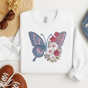 May include: White sweatshirt with a blue and purple butterfly embroidered on the front. The butterfly is surrounded by pink, white, and purple flowers.