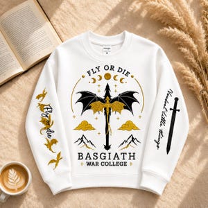 Pode incluir: Camisola branca com um desenho de drag&atilde;o e os textos "FLY OR DIE" e "BASGIATH + WAR COLLEGE +". O design inclui um drag&atilde;o, luas, nuvens e montanhas em preto e dourado. A manga tem os textos "Fly or die" e "Voudost titlle things".