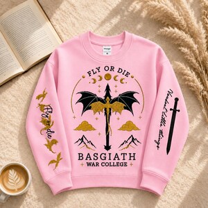 Pode incluir: Camisola rosa com um design de drag&atilde;o preto e detalhes dourados. Apresenta as frases "FLY OR DIE" e "BASGIATH + WAR COLLEGE +". As mangas t&ecirc;m texto e ilustra&ccedil;&otilde;es adicionais.