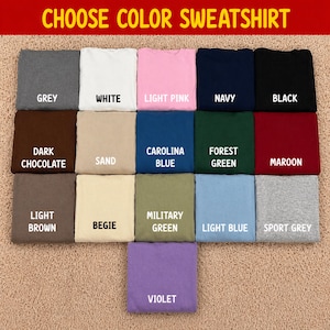 Pode incluir: Uma sele&ccedil;&atilde;o de camisolas dobradas em v&aacute;rias cores, incluindo cinzento, branco, rosa, azul marinho e preto. Cores adicionais s&atilde;o chocolate escuro, areia, azul Carolina, verde floresta, bord&ocirc;, castanho claro, bege, verde militar, azul claro, cinza desportivo e violeta. O texto no topo diz "CHOOSE COLOR SWEATSHIRT."