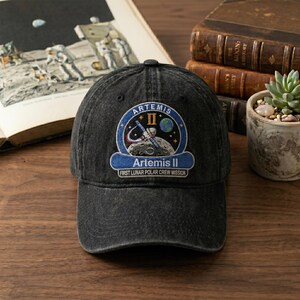 May include: A black baseball cap featuring an embroidered patch with the Artemis II mission logo. The patch includes the text "Artemis II" and "First Lunar Polar Crew Mission" with a design of the moon, Earth, and a rocket. The cap is sitting on a wooden surface.