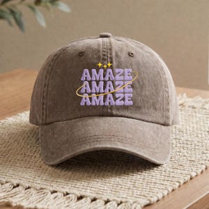 May include: A brown baseball cap with the word "AMAZE" in purple, stacked three times, with a gold swirl and stars. The cap is made of a soft material and has a curved brim.