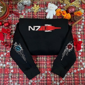 Peut inclure: Sweat-shirt noir avec le texte "NZ" en blanc et un graphisme rouge et blanc. Les manches présentent des graphismes blancs et rouges. Le sweat-shirt est présenté sur une surface à carreaux rouge et blanc avec des décorations de Noël.