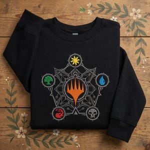 May include: A black long-sleeve sweatshirt featuring a central embroidered design of the Magic: The Gathering logo in orange, surrounded by a geometric star pattern and five circular icons representing the game's mana colors in yellow, green, blue, red, and white.