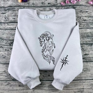 Puede incluir: Sudadera gris claro con un diseño negro de una criatura parecida a un lobo, una espada y un diseño de sol en la manga. Tiene cuello redondo y mangas largas.