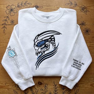 Puede incluir: Sudadera blanca con un gráfico detallado en negro y azul de la cara de un personaje en la parte delantera. La manga izquierda tiene un diseño azul y blanco, y la manga derecha tiene el texto "THERE IS NO SHEPARD WITHOUT VAKARIAN".