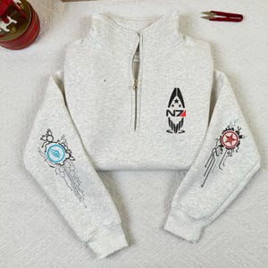 Op de afbeelding: Een lichtgrijs sweatshirt met een kwartrits en een hoge kraag. Het sweatshirt heeft een zwart-rode afbeelding op de borst en gedetailleerde ontwerpen op de mouwen. De mouwen hebben een blauw en rood grafisch ontwerp. De rits is zilver.