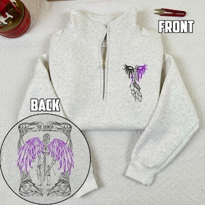 Puede incluir: Sudadera gris claro con cremallera de cuarto de cierre y un diseño gráfico negro y morado en la parte delantera. La parte trasera de la sudadera presenta un diseño circular con el texto "THE DEMON OF VYRANTIUM" y una figura alada morada.