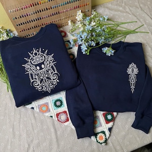 Puede incluir: Dos sudaderas azul marino de cuello redondo con bordados de líneas blancas. La sudadera de la izquierda muestra un diseño de personaje grande e intrincado con patrones circulares y de remolinos. La de la derecha presenta un emblema vertical simétrico.
