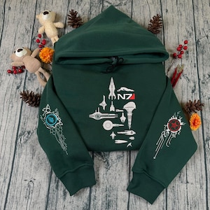 Puede incluir: Sudadera con capucha verde oscuro con diseños gráficos blancos y el texto "N7". La sudadera presenta varias ilustraciones de naves espaciales en la parte delantera y diseños detallados en las mangas. La sudadera con capucha se muestra sobre una superficie de madera.
