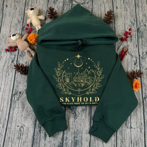 Puede incluir: Una sudadera con capucha verde oscuro con un gráfico dorado de un castillo, montañas y elementos celestiales. El texto "SKYHOLD" y "THE PLACE WHERE THE SKY IS KEPT" están debajo. La sudadera tiene una capucha con cordón.