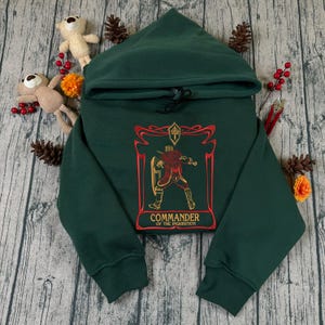 May include: Dark green hoodie with a drawstring hood. The front has an embroidered design of a warrior with a shield and the text "COMMANDER OF THE INQUISITION" in red and gold. The hoodie is made of a soft material.