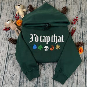 May include: A dark green hooded sweatshirt featuring the white text I'd tap that printed across the chest. Below the text are five small colorful icons representing a blue water drop, a green tree, a white skull, a red flame, and a yellow sun.