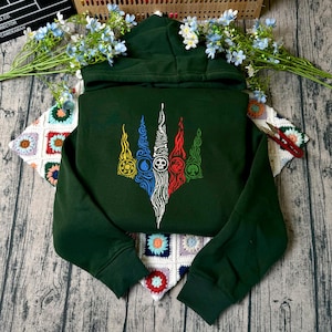 Könnte beinhalten: Ein dunkelgrüner Kapuzenpullover mit einem zentralen Grafikdesign, das aus fünf vertikalen, flammenartigen Formen in Gelb, Blau, Weiß, Rot und Grün besteht. Jede Form enthält ein kreisförmiges Symbol für Sonne, Wasser, Totenkopf, Drache und Baum.