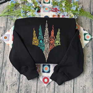 May include: A black long-sleeve sweatshirt featuring a central graphic print of five vertical, flame-like shapes in yellow, teal, beige, orange, and green. Each shape contains a circular icon representing sun, water, skull, dragon, and tree symbols.