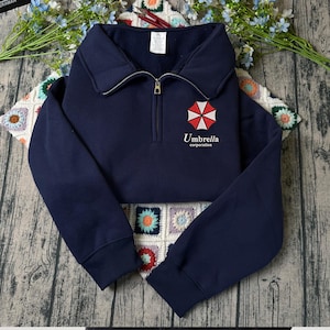 Peut inclure: Un sweat-shirt bleu marine à fermeture éclair quart, doté d'un col rabattu et de manches longues à poignets côtelés. La poitrine présente un motif de parapluie rouge et blanc au-dessus du texte Umbrella corporation en lettres blanches.