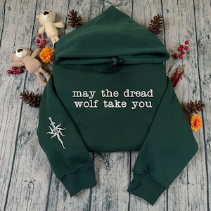 Puede incluir: Una sudadera con capucha verde oscuro con el texto blanco "may the dread wolf take you". Tiene una capucha con cordón y un gráfico blanco en la manga izquierda. La sudadera se presenta sobre una superficie de madera, decorada con pequeños elementos.