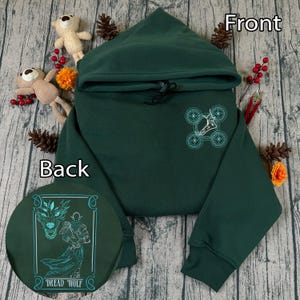 Puede incluir: Una sudadera con capucha verde oscuro con diseños color teal en la parte delantera y trasera. La parte delantera presenta un diseño teal, y la trasera muestra una ilustración con las palabras "Dread Wolf". La sudadera tiene capucha y mangas largas.