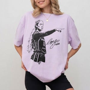 May include: A light purple short-sleeve t-shirt featuring a black graphic illustration of a basketball player pointing forward. The player wears a jersey with the number 8 and stars on the side. A black signature is printed next to the player.