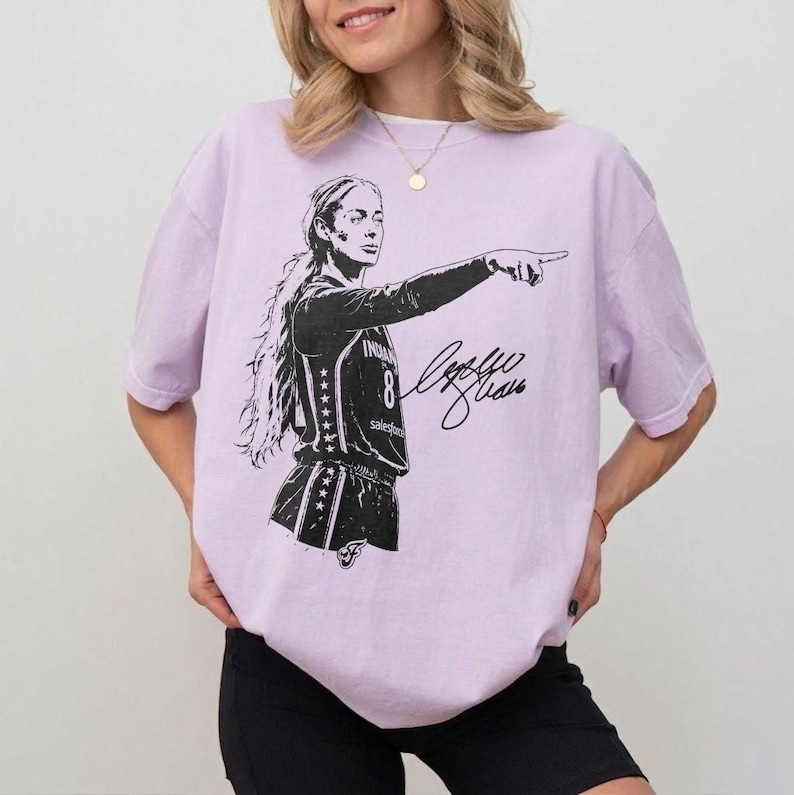 May include: A light purple short-sleeve t-shirt featuring a black graphic illustration of a basketball player pointing forward. The player wears a jersey with the number 8 and stars on the side. A black signature is printed next to the player.