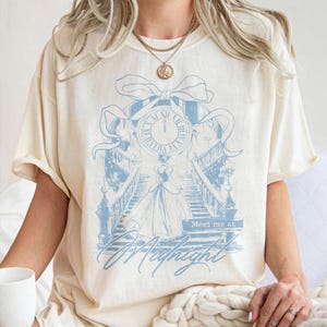Pu&ograve; includere: T-shirt color crema con un'illustrazione azzurra di un orologio, una scala e una donna in abito. Il testo "Meet me at Midnight" &egrave; sotto l'illustrazione. La maglietta ha maniche corte e una vestibilit&agrave; ampia.