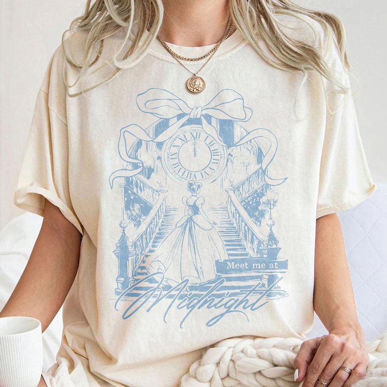 Pu&ograve; includere: T-shirt color crema con un'illustrazione azzurra di un orologio, una scala e una donna in abito. Il testo "Meet me at Midnight" &egrave; sotto l'illustrazione. La maglietta ha maniche corte e una vestibilit&agrave; ampia.