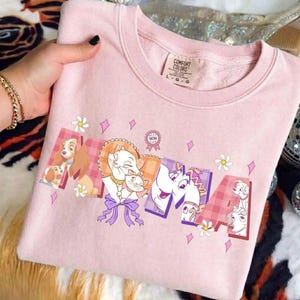 Puede incluir: Una sudadera de cuello redondo rosa claro con la palabra MAMA en letras grandes. Las letras están rellenas con ilustraciones de personajes de Disney, incluyendo a Lady, Marie, Duchess y la Sra. Potts. Pequeñas flores blancas y diamantes rosas rodean el texto.
