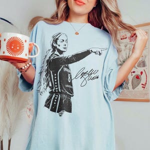 May include: A light blue t-shirt featuring a black graphic of a basketball player wearing a jersey with the number 8 and the text Indiana, accompanied by a signature. The shirt is paired with a white mug displaying a large orange sun graphic.