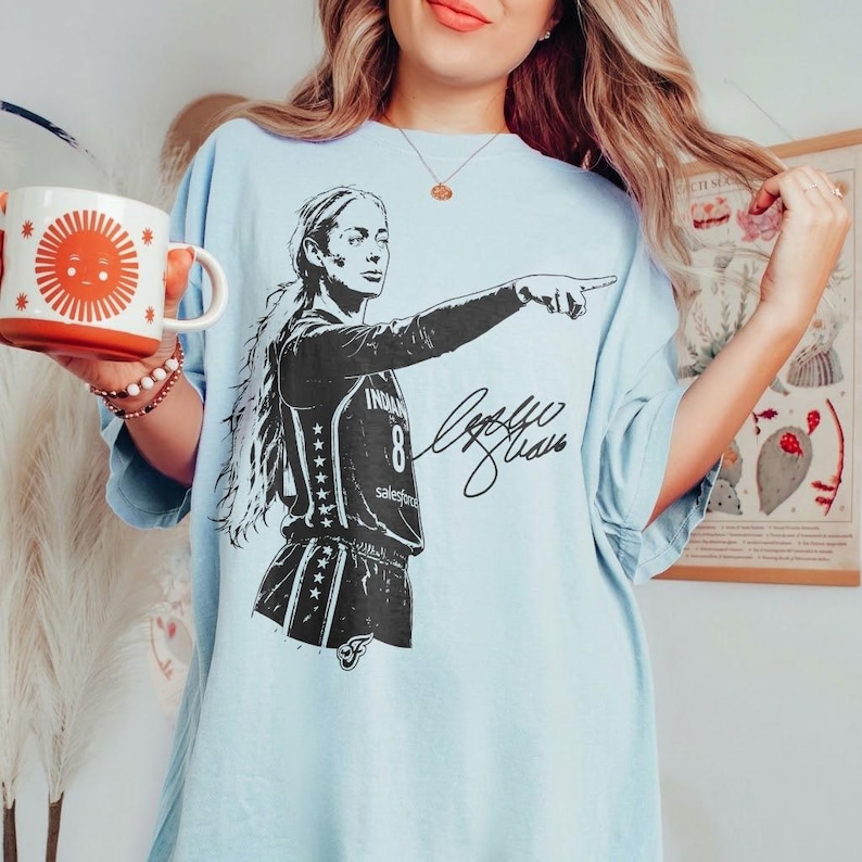 May include: A light blue t-shirt featuring a black graphic of a basketball player wearing a jersey with the number 8 and the text Indiana, accompanied by a signature. The shirt is paired with a white mug displaying a large orange sun graphic.