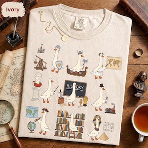 Peut inclure: Un t-shirt ivoire avec une impression graphique représentant diverses oies blanches vêtues de costumes historiques, notamment un Viking, un Romain et un pharaon égyptien. Le motif inclut des illustrations d'un château, d'une carte, d'un tableau noir avec le texte HISTORY, d'une bibliothèque et d'une usine, le tout dans un style dessin animé.