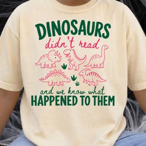 Peut inclure: Un t-shirt à manches courtes de couleur crème avec une impression graphique comportant du texte vert foncé et rose qui dit : DINOSAURS didn't read and we know what HAPPENED TO THEM. Le motif inclut cinq illustrations au trait rose de dinosaures.