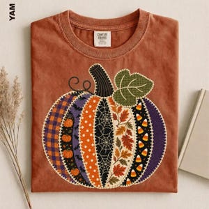 Peut inclure: Un t-shirt couleur rouille arborant un grand motif central de citrouille composé de segments verticaux style patchwork. Les segments présentent divers motifs, notamment des carreaux, des pois, des toiles d'araignée, des feuilles d'automne et des bonbons.